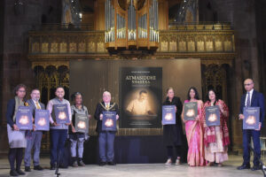 a group of people stand around a book poster in manchester cathedral at gandhi's birthday event. the book poster announces the english translation of atmasiddhi shastra